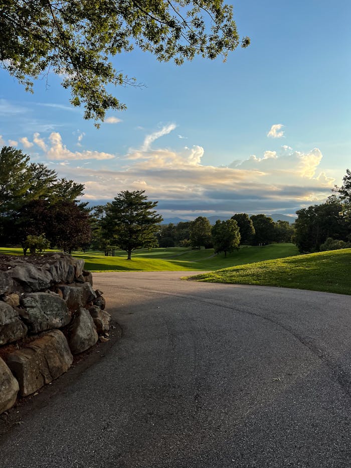 A peaceful park scene at sunset featuring a winding road, lush trees, and a dramatic sky.